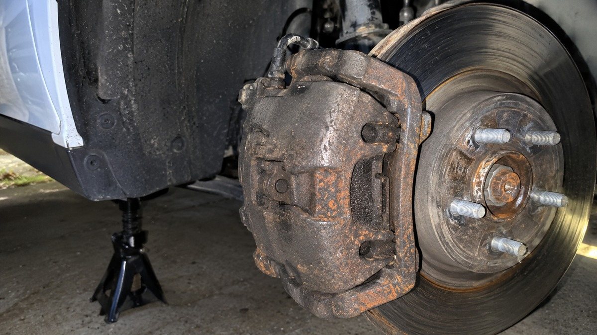 Dramatized scene: a gray stock brake caliper crusted in brake dust and rust with the wheel removed